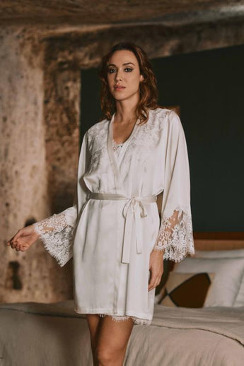 Model wearing the Marjolaine Tess Short Silk Robe in ivory featuring smooth silk satin, floral lace cuffs, a wrap-front design, long sleeves, and a self-tie waist belt.
